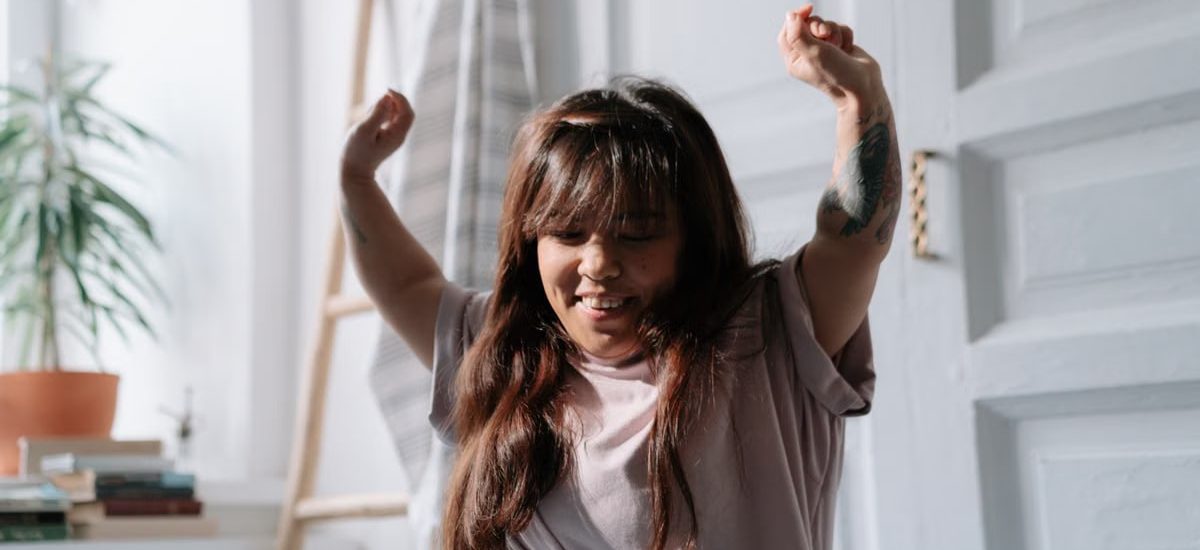 A relaxed Asian woman wakes up and stretches in a sunlit bedroom, enjoying the morning light.